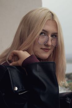 Portrait of a blonde woman wearing eyeglasses, smiling confidently outdoors in a leather jacket.