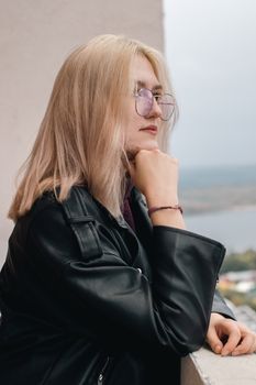 Pensive woman in a black leather jacket with blonde hair looking afar outdoors.