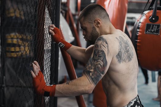 A tattooed boxer taking a break while leaning against a gym fence with red boxing gloves.