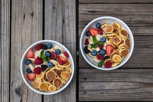 Top view of two bowls with mini pancakes and fresh fruits on rustic wood table.