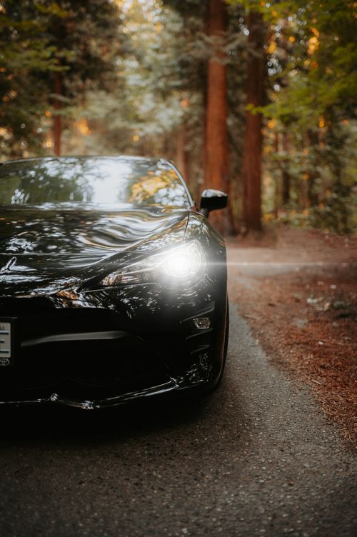 Free A sleek black sports car with headlights on, parked on a forest road, exuding a modern and fast vibe. Stock Photo