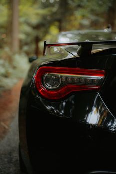 Close-up view of a sleek black car's taillight in a wooded area, highlighting design and nature contrast.