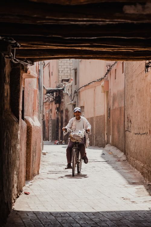 Free A man rides a bicycle through a sunlit, narrow alleyway of an urban setting, capturing everyday life. Stock Photo