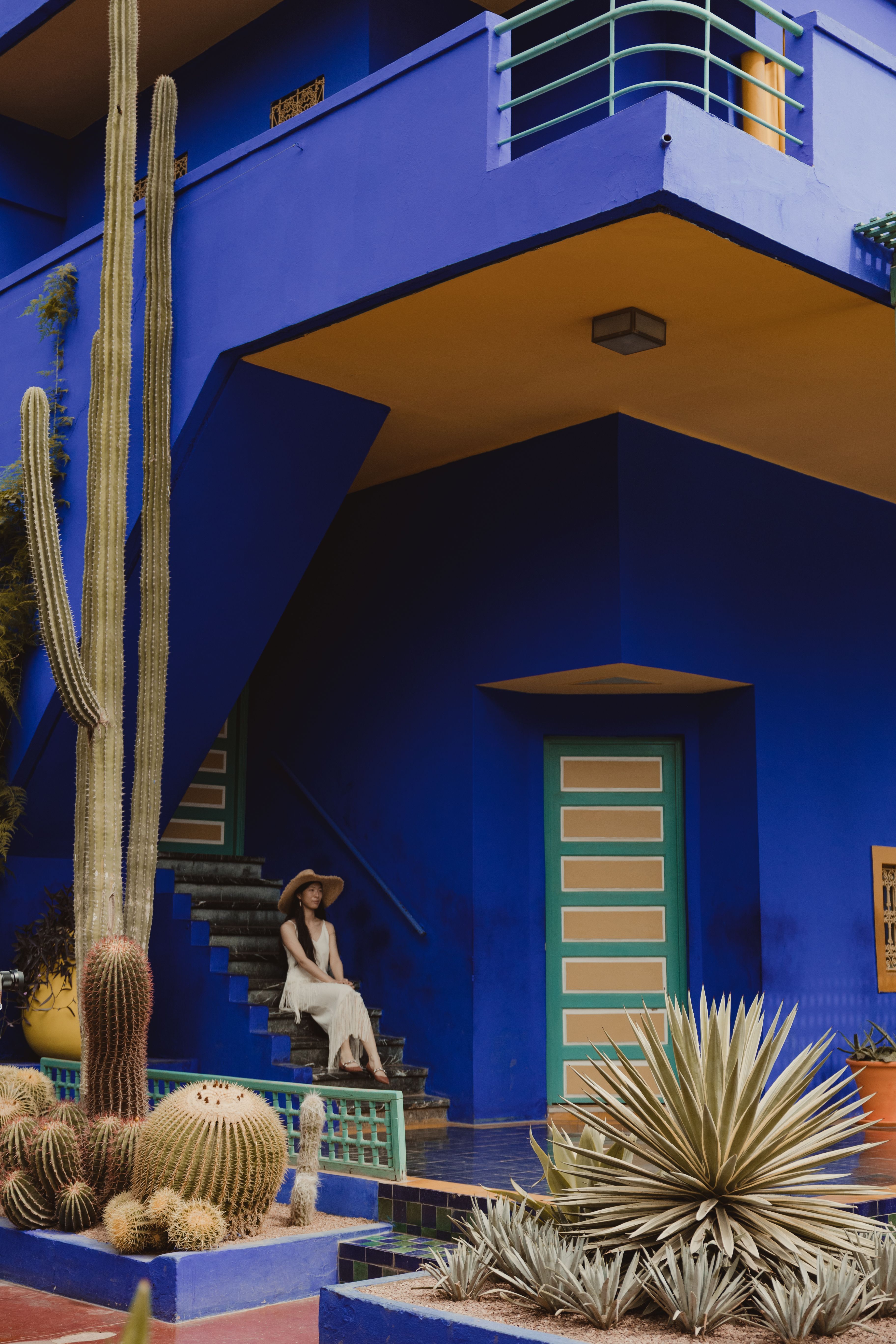 Woman in a hat sitting on steps of colorful building with cacti.