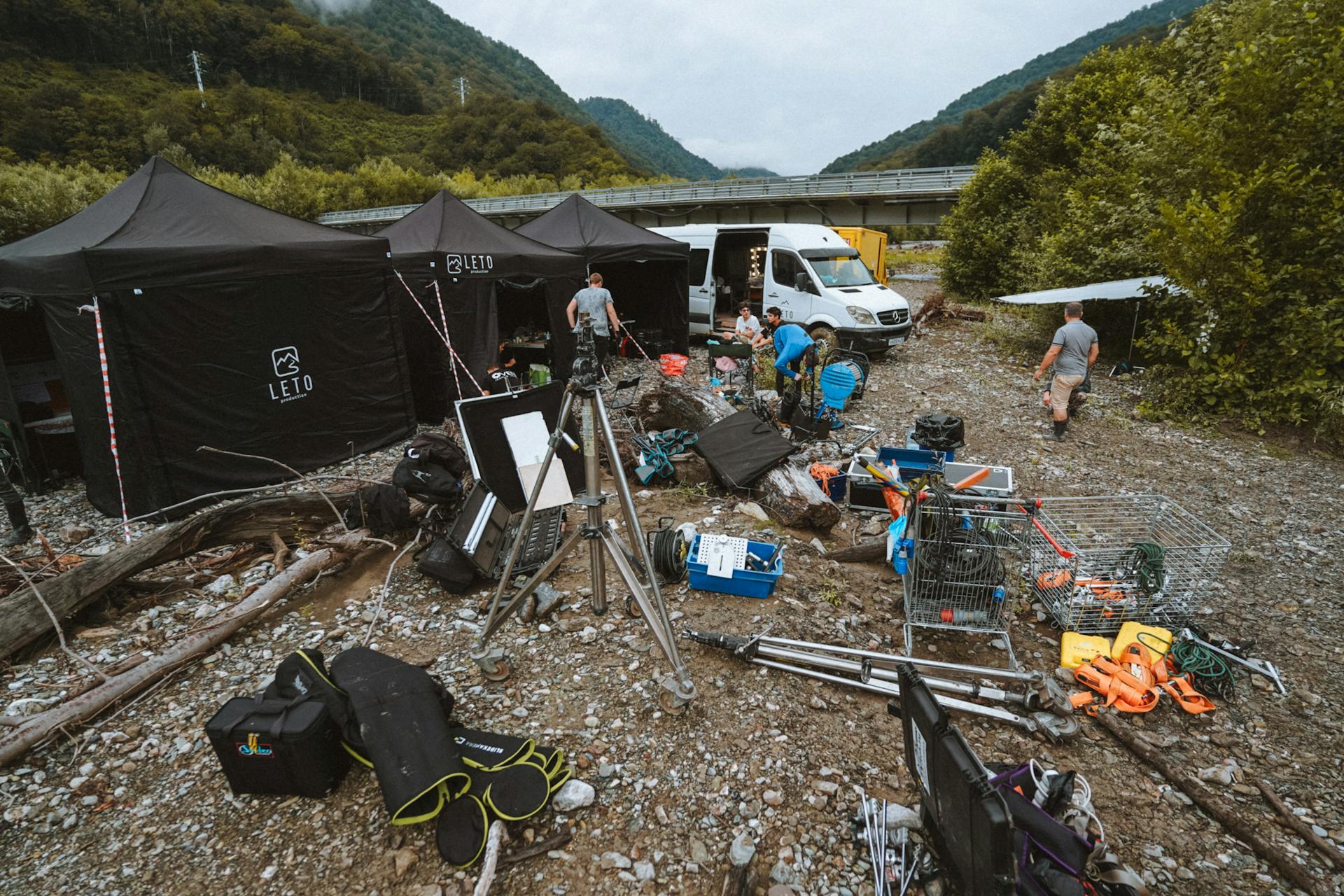 Film crew setting up outdoor equipment for a shoot near mountains with tents and vehicles.