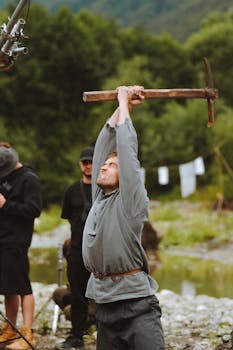 Actor on a movie set outdoors holding a pickaxe, surrounded by crew in a mountainous area.