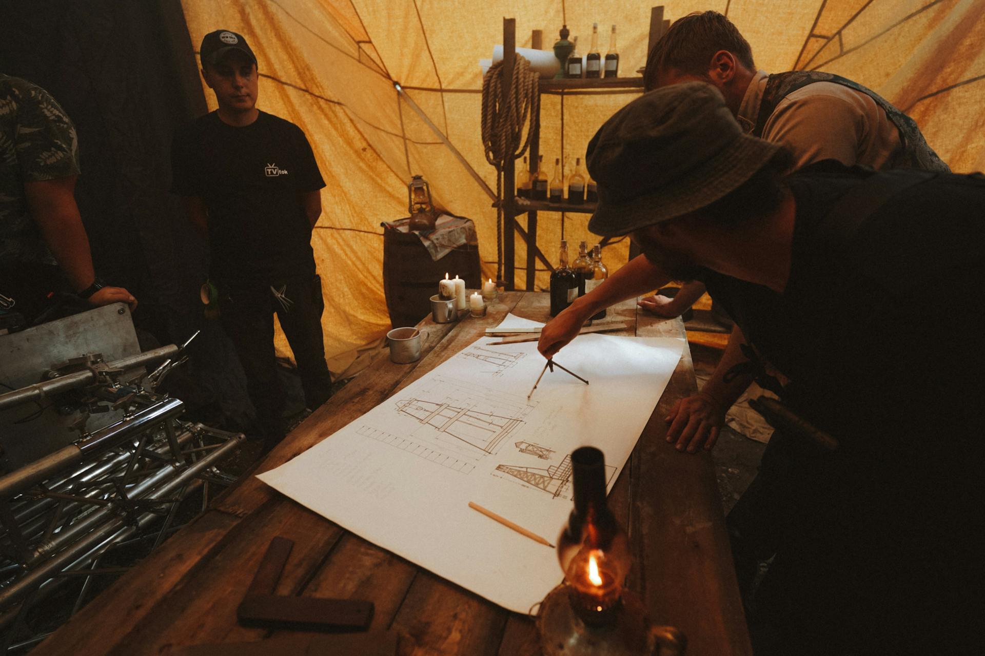 A group discussing architectural plans on a wooden table in dimly lit tent environment.