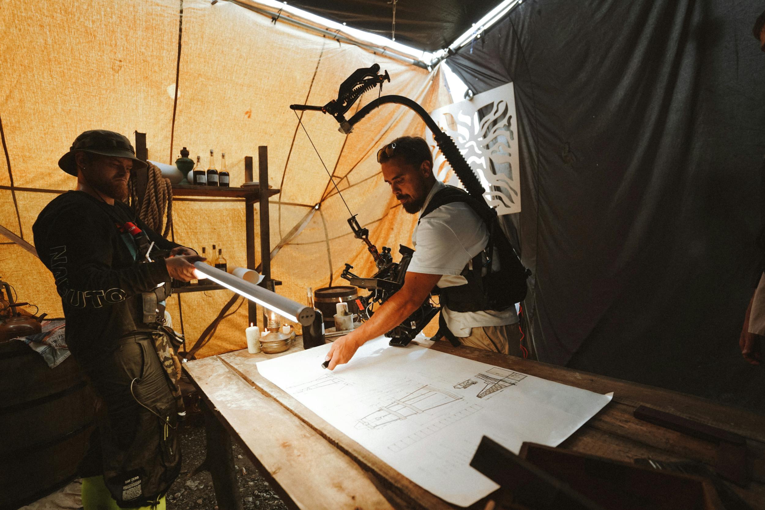 Two engineers use detailed plans inside a tented workshop, focusing on construction or design projects.