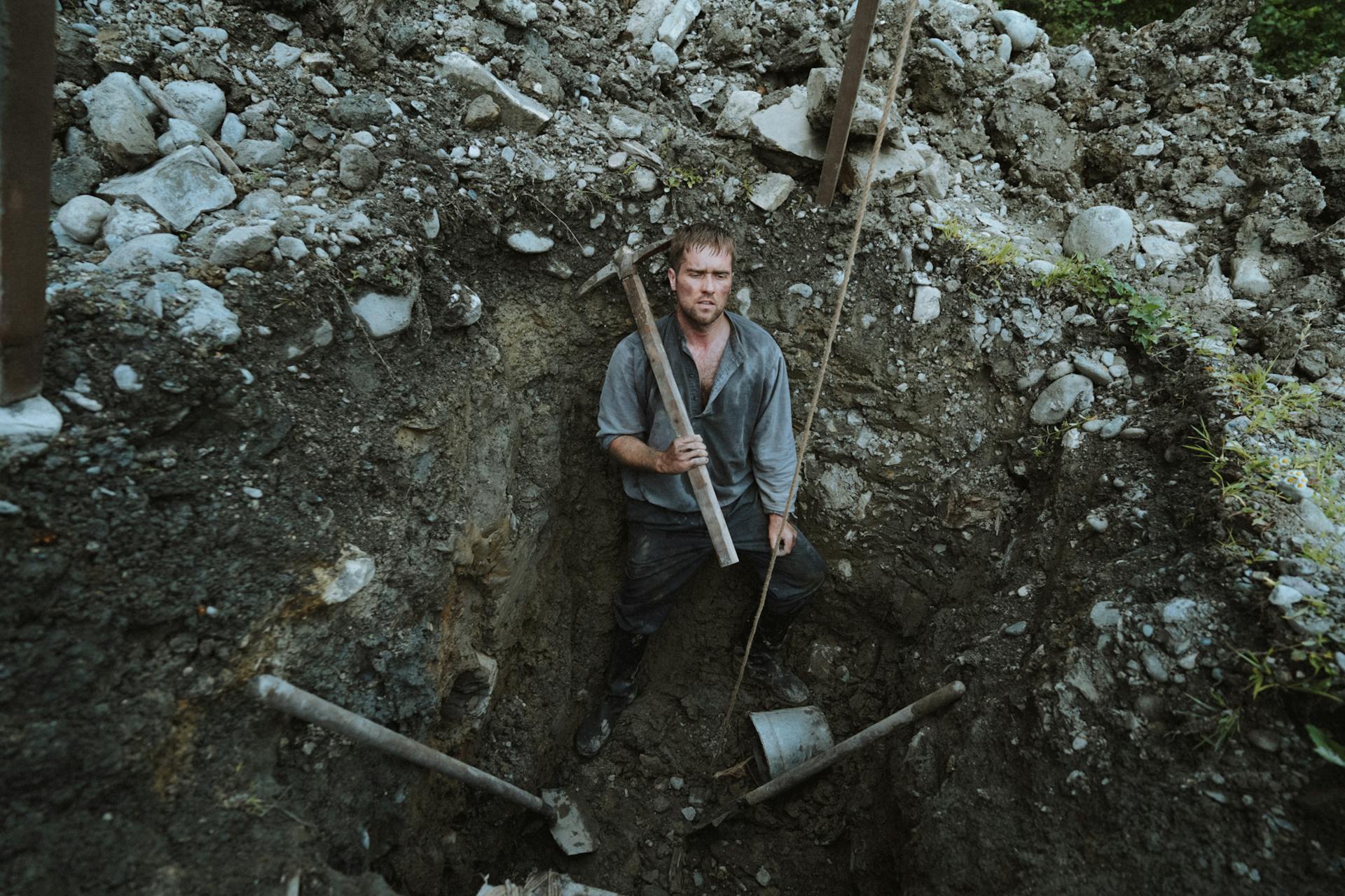 A rugged miner stands in a deep excavation holding a pickax, surrounded by rocky terrain.