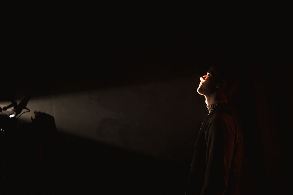 A man stands in darkness with a spotlight illuminating his face, creating a dramatic effect.