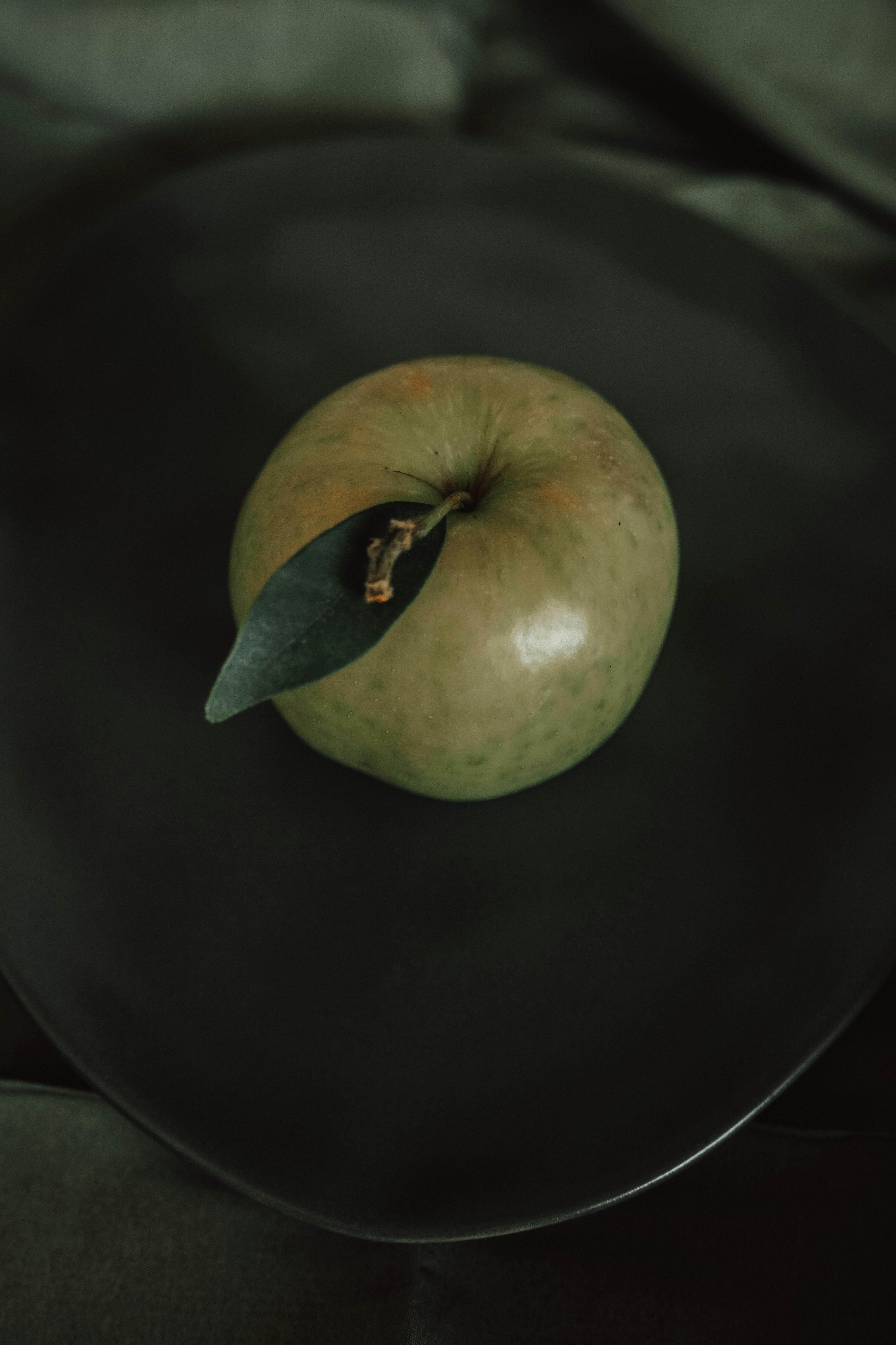 Artistic photo of a green apple with leaf on a dark plate, capturing a moody aesthetic.