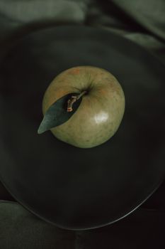 Artistic photo of a green apple with leaf on a dark plate, capturing a moody aesthetic.
