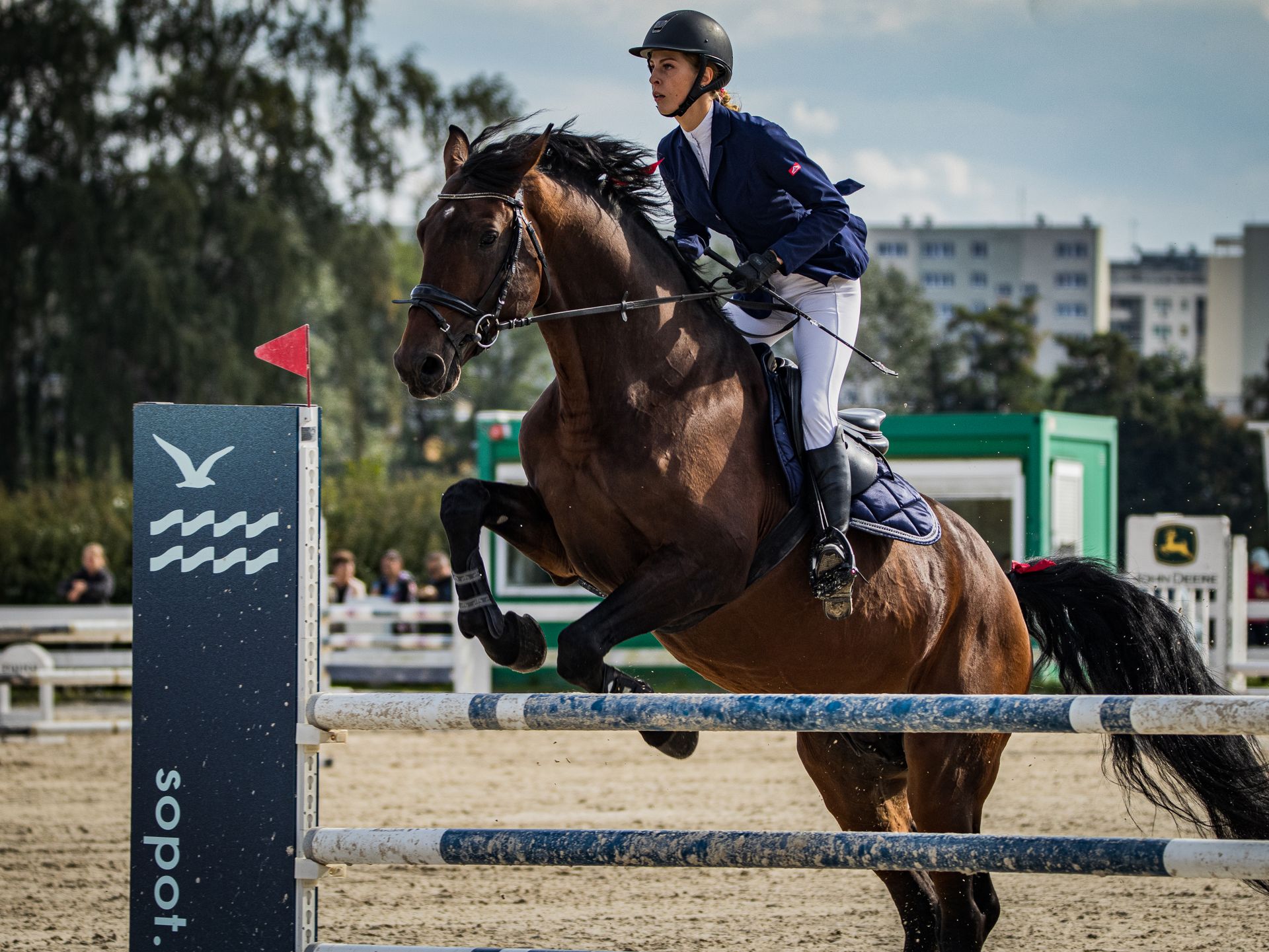 A female rider and brown horse gracefully navigate an equestrian jump in a sunny outdoor arena.