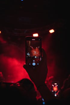 A close-up of a cellphone recording a vibrant concert with red stage lights.