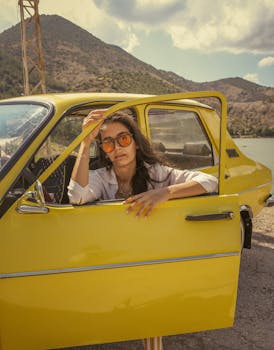 Woman in sunglasses leaning out of a yellow car door, set against a scenic mountain backdrop.