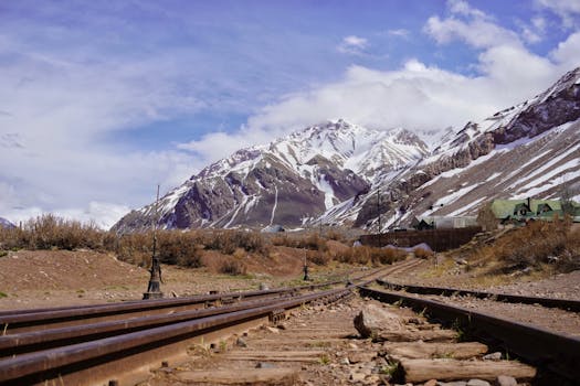 A stunning view of the snow-capped Andes mountains and a railroad track in Argentina under a cloudy sky.