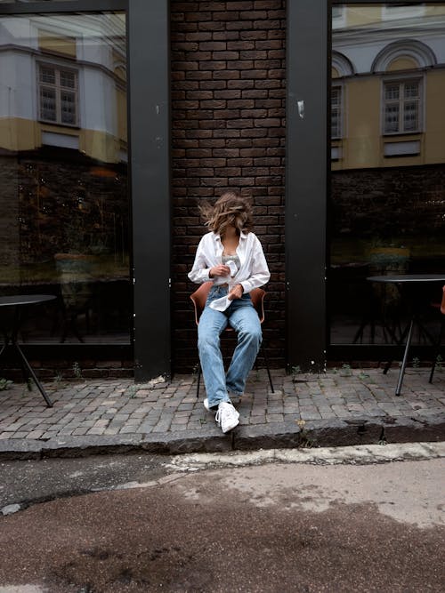 A Woman in White Long Sleeves and Denim Jeans Sitting on a Chair Near Brick Wall