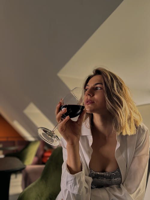 A Woman in White Long Sleeves Holding Goblet Glass with Wine while Looking Afar