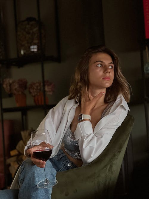 A Woman in a White Long Sleeved Shirt Holding a Glass of Wine while Sitting on a Chair