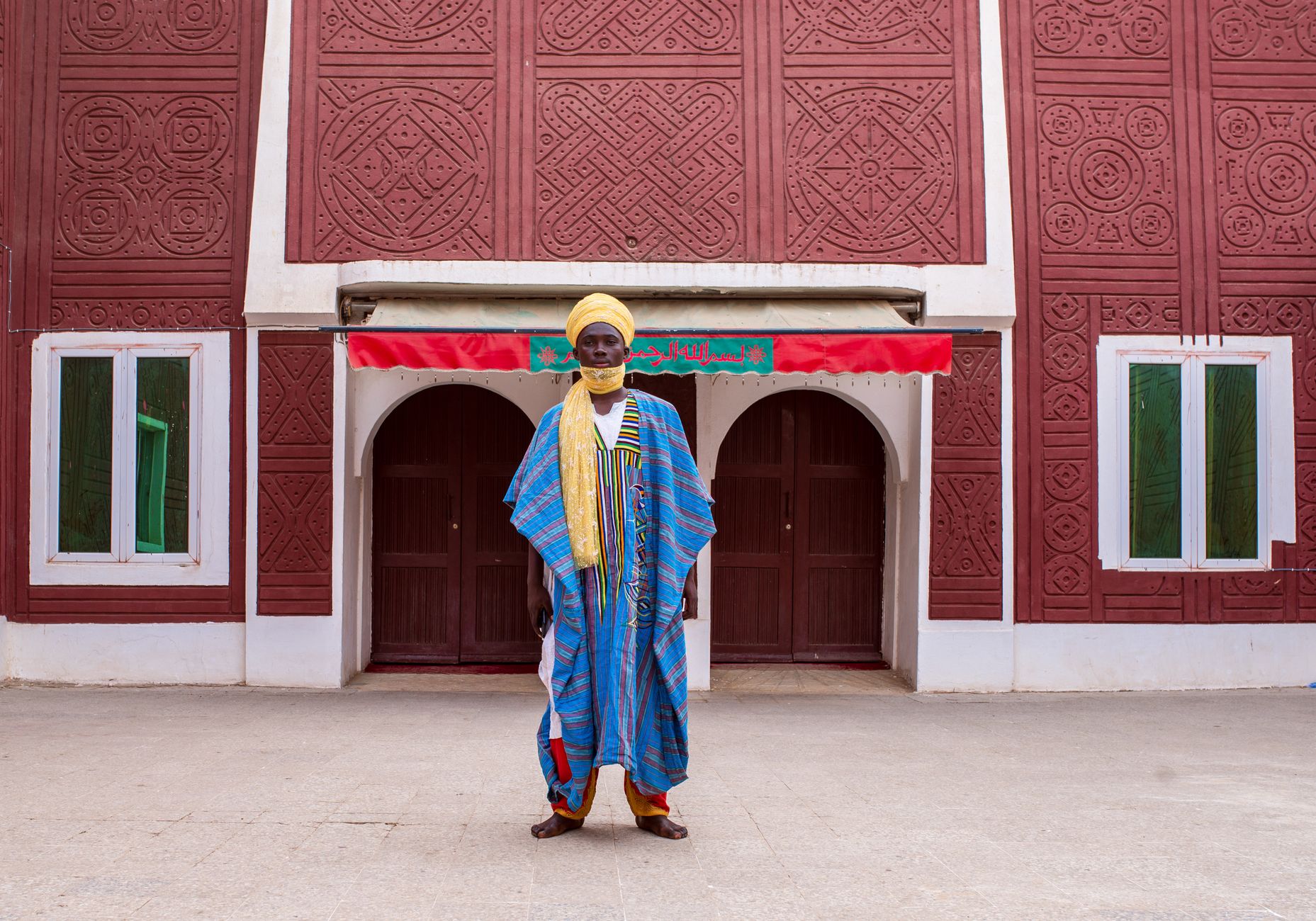 A man in traditional attire stands before a decorated building in Kano, Nigeria.