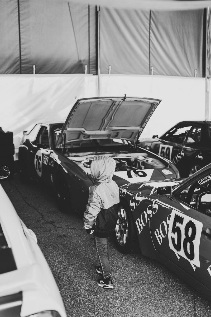 A child standing amidst racing cars in a garage, captured in black and white.