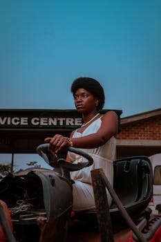Determined black woman in white dress driving a tractor near a service center.