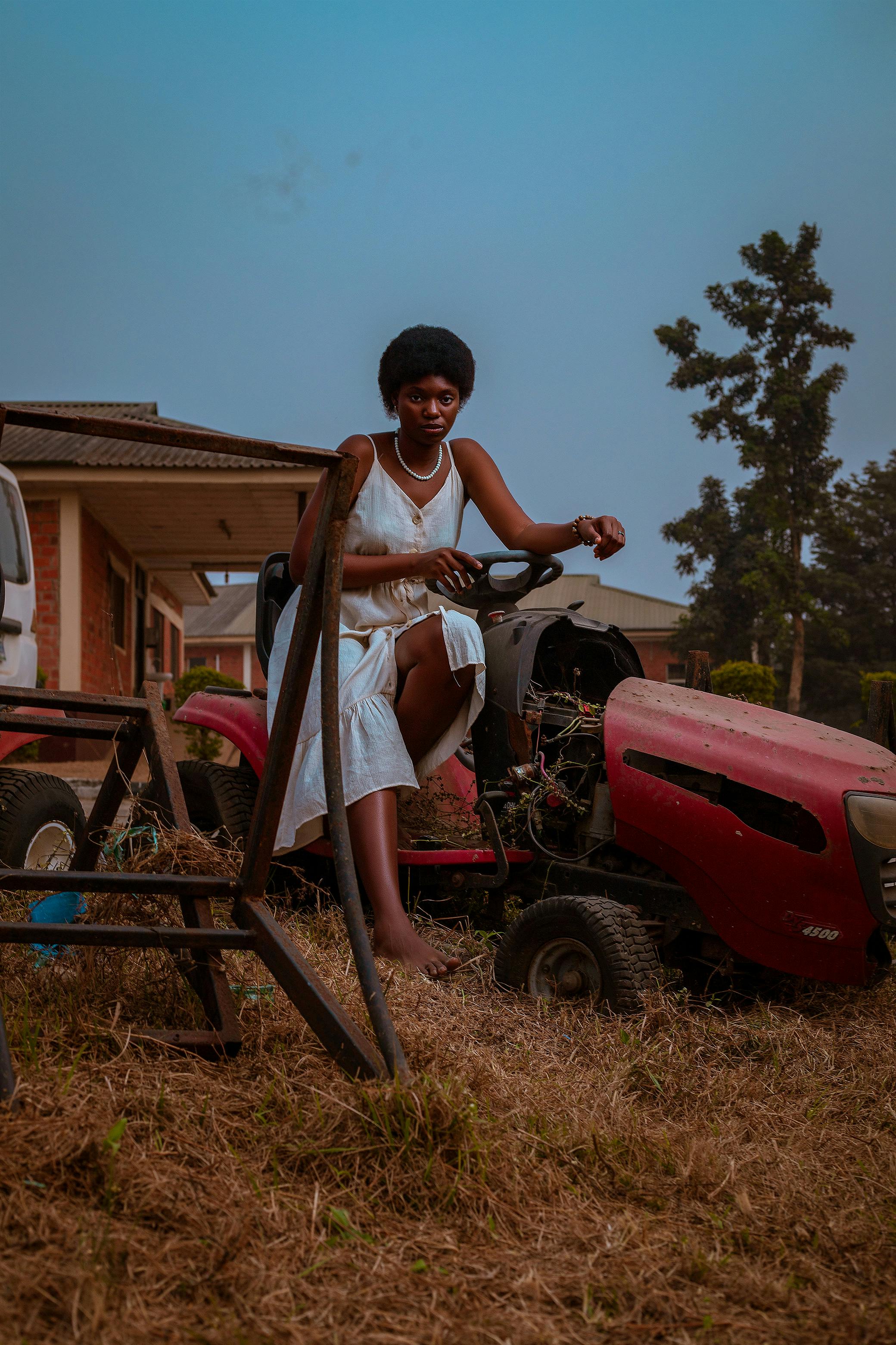 Young African woman in white dress sitting on a lawn mower in a rural outdoor setting.