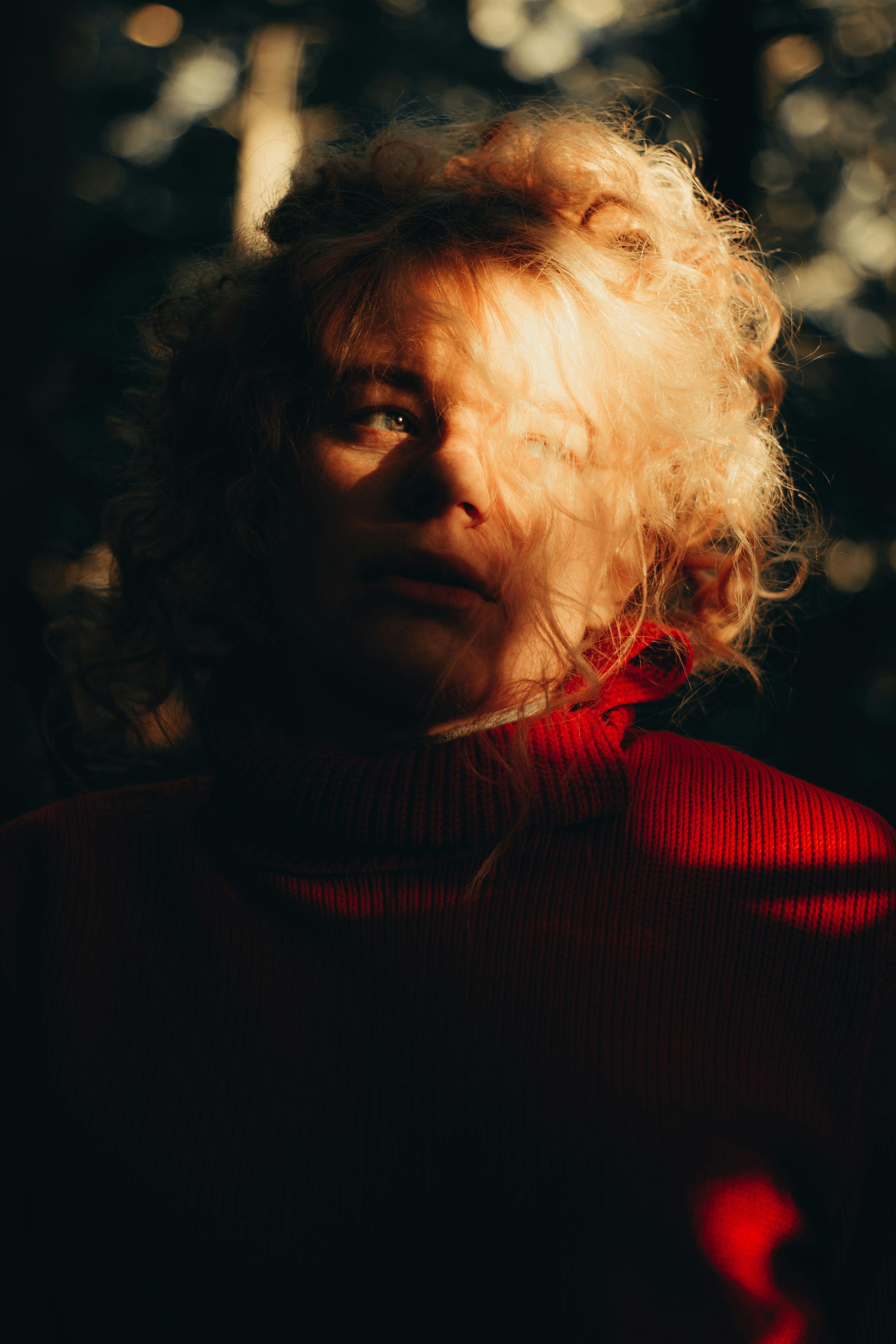 Moody portrait of a young woman with blonde hair in a red sweater, illuminated by sunlight in a forest.