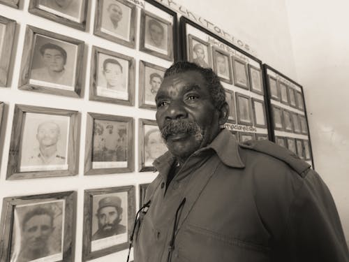 Grayscale Photography of an Elderly Man Standing Near Wall with Hanging Picture Frames