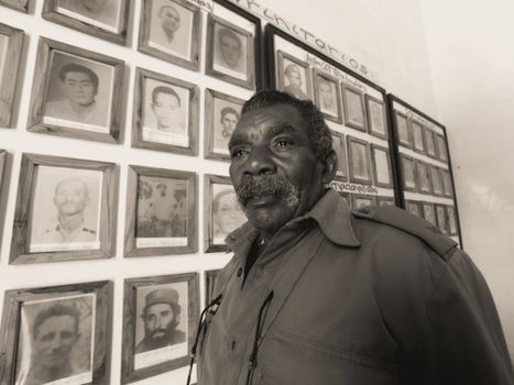Senior man standing by a wall of framed historical photos, in a monochrome setting.