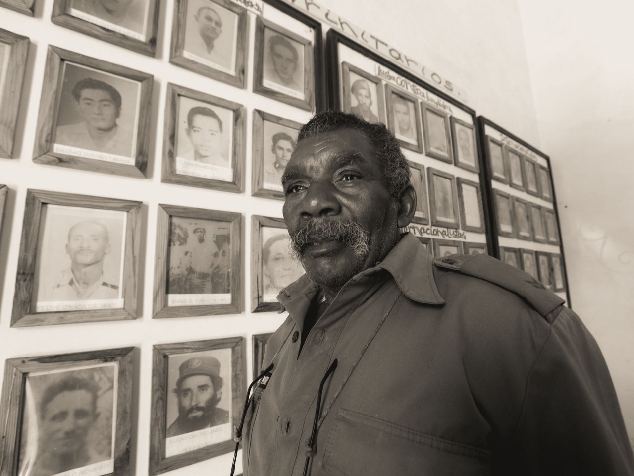 senior man standing by a wall of framed historical photos in a monochr