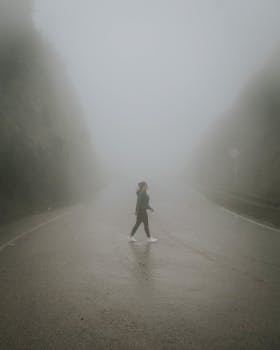 A solitary person walks on a foggy road surrounded by dense mist, evoking a mysterious atmosphere.
