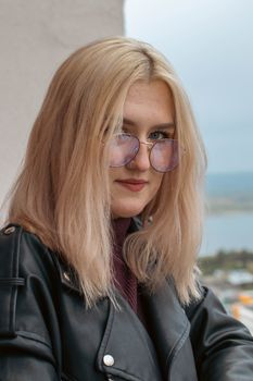 Fashionable woman with blonde hair and glasses poses stylishly in a black leather jacket.