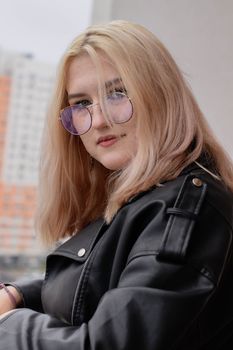 Stylish woman in a black leather jacket and glasses gazing confidently over her shoulder outdoors.