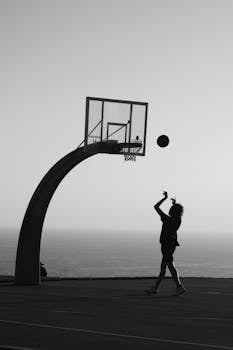 Silhouette of a person shooting a basketball on an outdoor court by the sea.