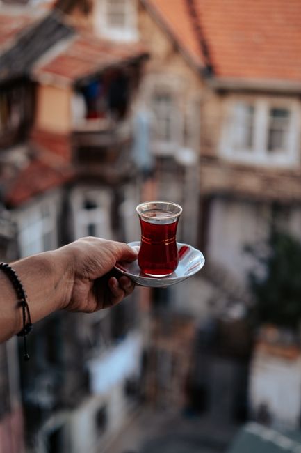 A hand holding a glass of traditional Turkish tea with a blurred cityscape background.