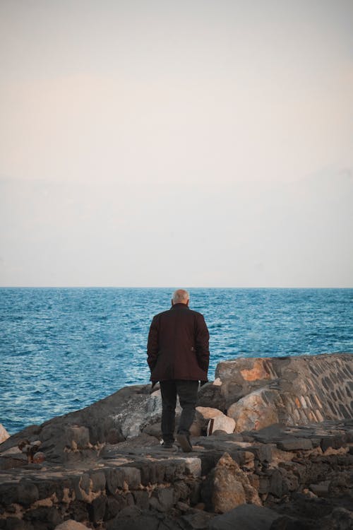 A Back View of a Man in Black Suit Standing on the Rock Near the Sea
