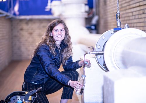 Free Smiling woman fills a bottle from an industrial pipe in a factory setting. Stock Photo