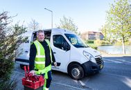 Smiling worker in reflective vest stands by a van holding tools.