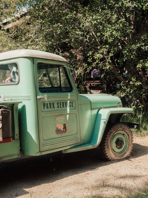 A classic green park service truck parked on a sunny dirt road surrounded by lush trees.