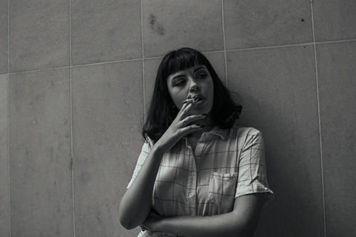 Free A woman in a checkered shirt leans against a tiled wall, smoking a cigarette. Stock Photo