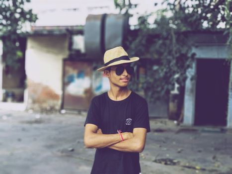 Young man in sunglasses and hat poses with arms crossed outdoors.