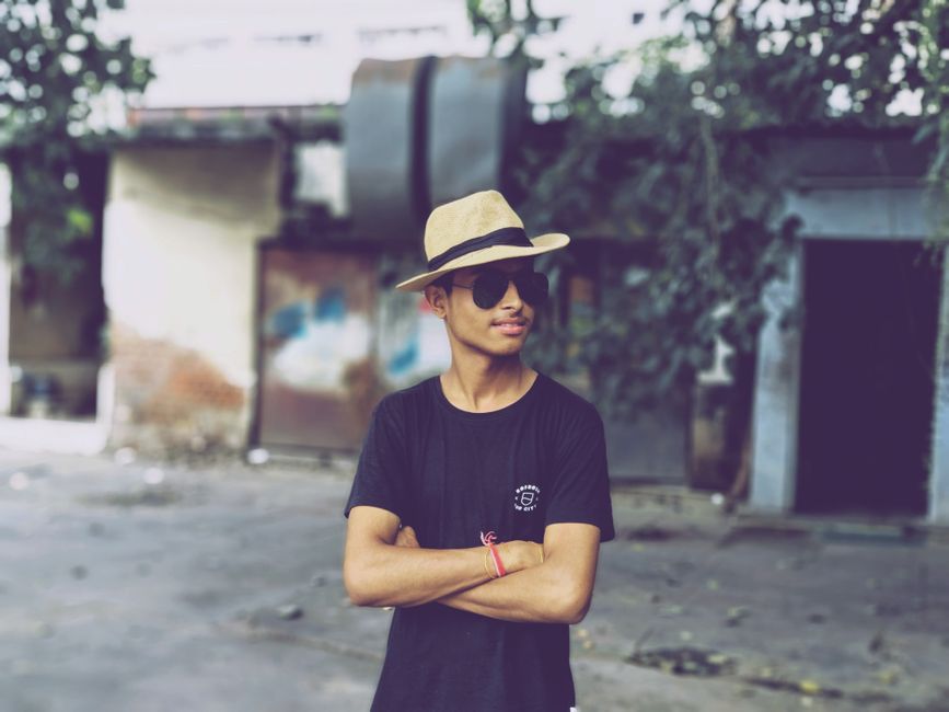 Young man in sunglasses and hat poses with arms crossed outdoors.