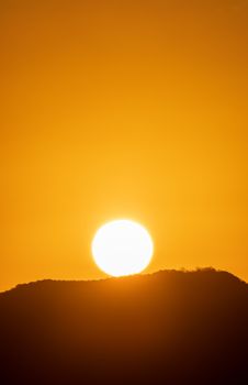 Silhouetted mountain peaks against a vivid orange sunset sky with the sun setting on the horizon.