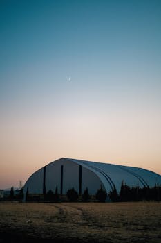 Serene dawn scene with a large hangar under a crescent moon in the countryside.