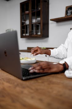 An individual multitasking with a laptop and food on a wooden table indoors.