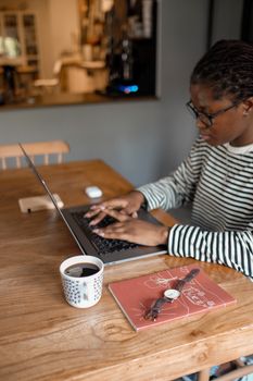 African American woman working remotely with laptop, coffee, and notebook in a cozy indoor setting.