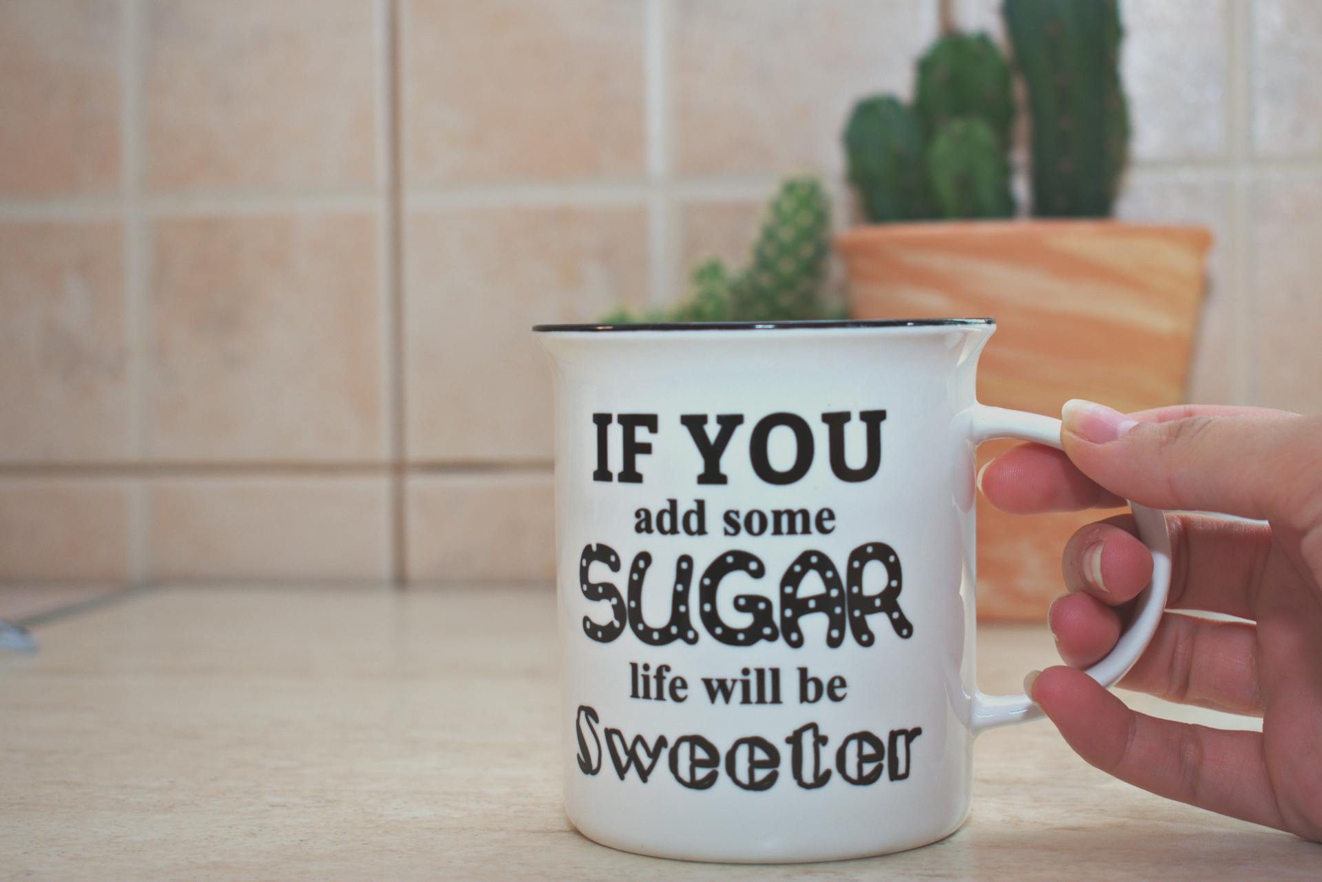 A hand holds a mug with a playful quote about sugar, set against a kitchen background.