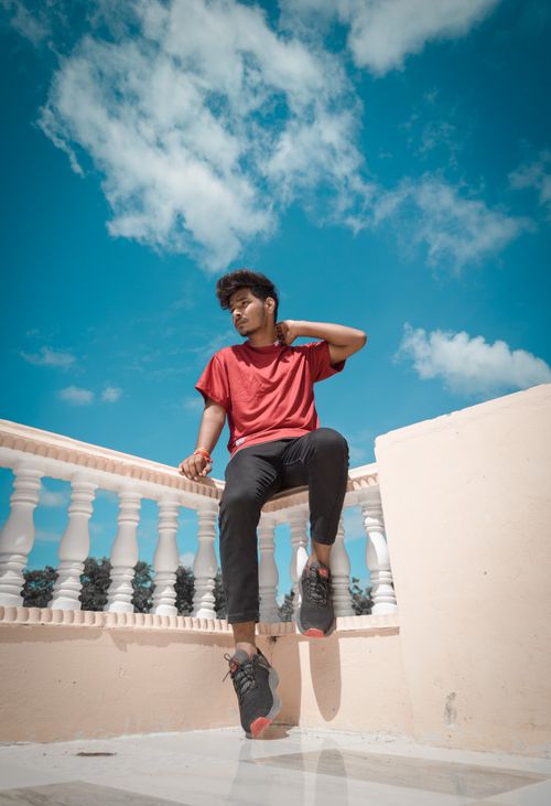 Free A stylish young man in a red t-shirt posing on a rooftop balcony under a bright blue sky. Stock Photo