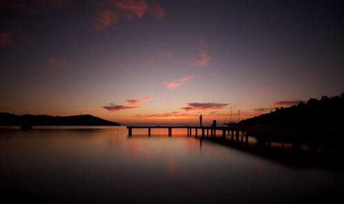 A Silhouette of a Person Standing on a Dock Near Body of Water During Sunset